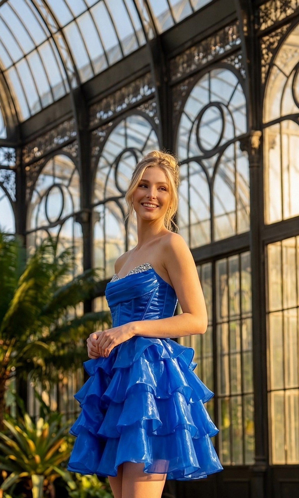 A woman wearing the Short Homecoming Dress: Juliet JT7134S, a strapless organza design with a ruffle A-line mini skirt, stands in a glass conservatory with arched windows and sunlight streaming in.